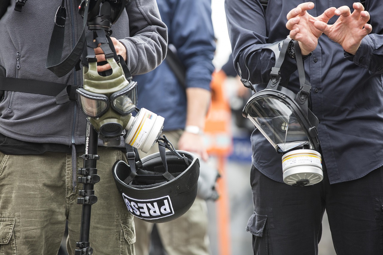 Dwaj dziennikarze w maskach gazowych i kaskach z napisem PRESS, przygotowani na niebezpieczne sytuacje.