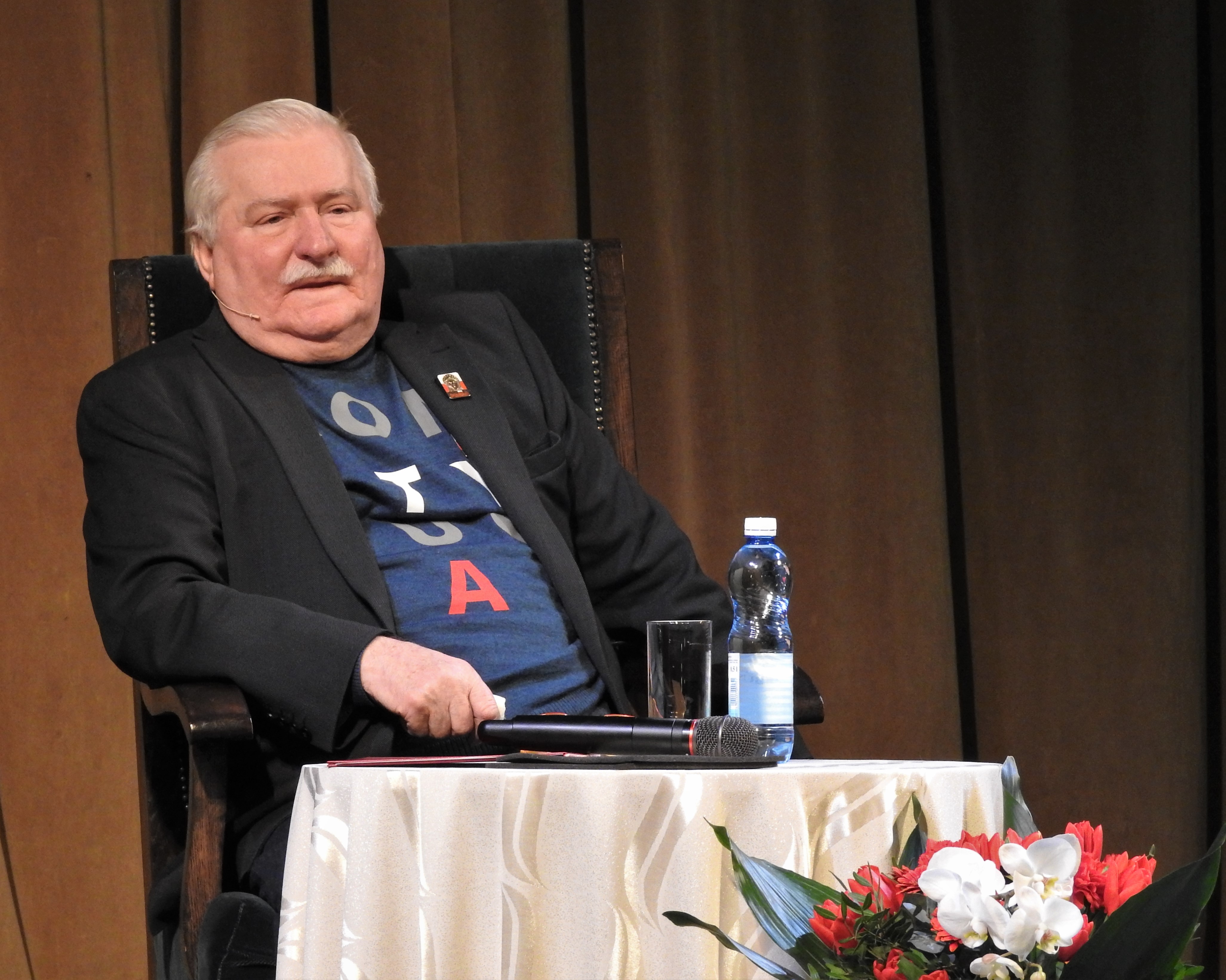 Lech Wałęsa w garniturze i koszulce z napisem "Solidarność" siedzi w fotelu, przed nim mikrofon i butelka wody.