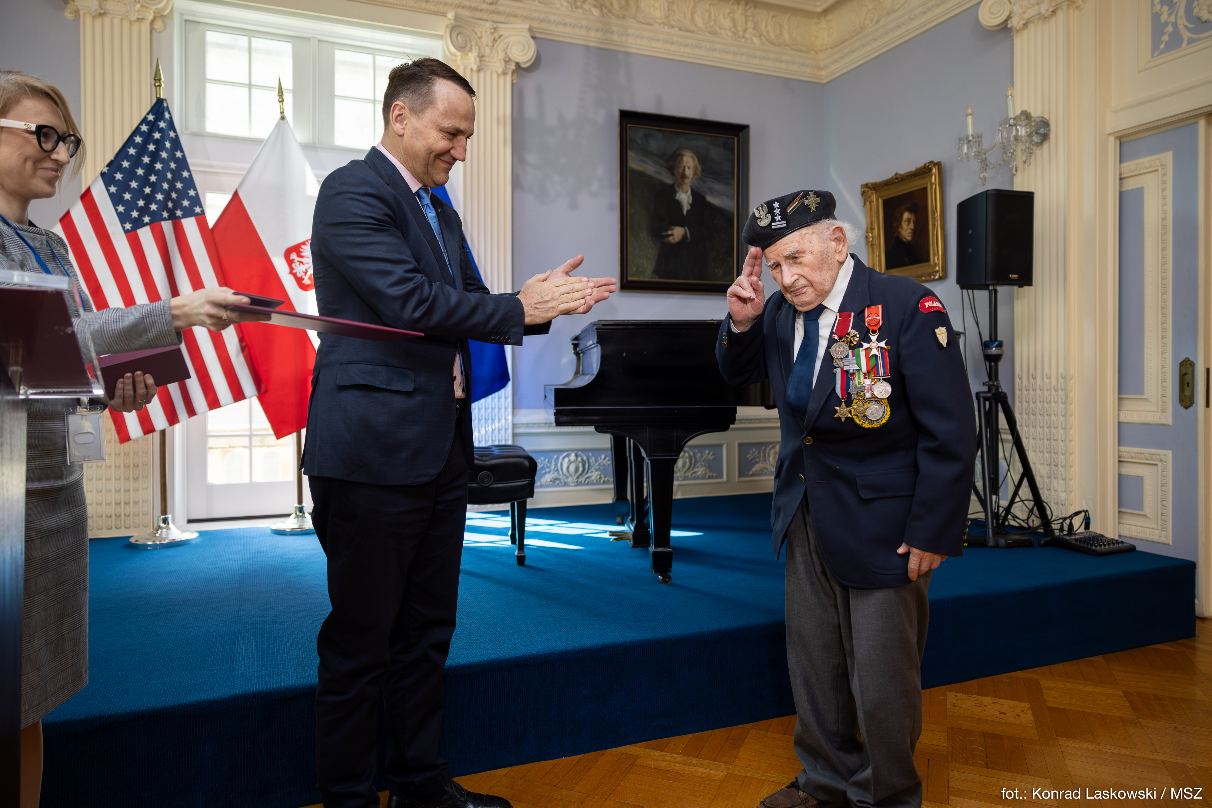 Minister Sikorski wręcza dokument weteranowi z odznaczeniami, w tle flagi Polski i USA.