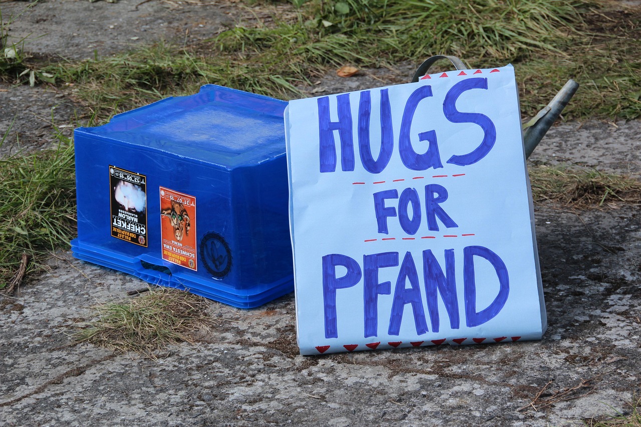 Niebieski pojemnik obok tablicy z napisem 'Hugs for Pfand' w kontekście systemu kaucyjnego na butelki PET.