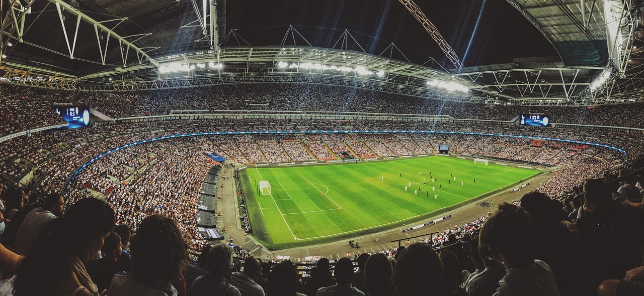 Stadion piłkarski z pełnymi trybunami i oświetloną murawą, sugerujący atmosferę meczu.