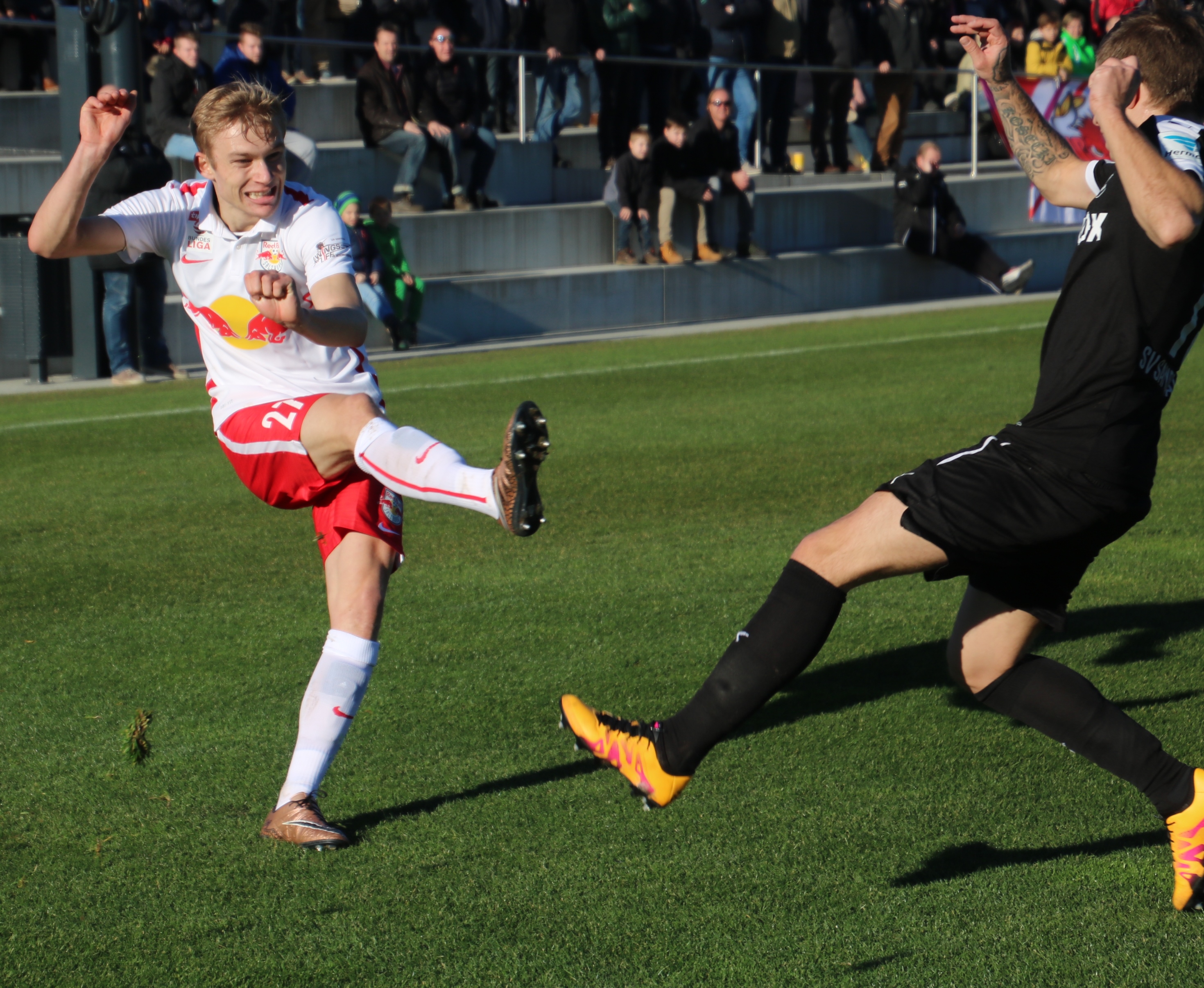 Piłkarz w białej koszulce z herbem Red Bull Salzburg kopie piłkę na zielonej murawie.