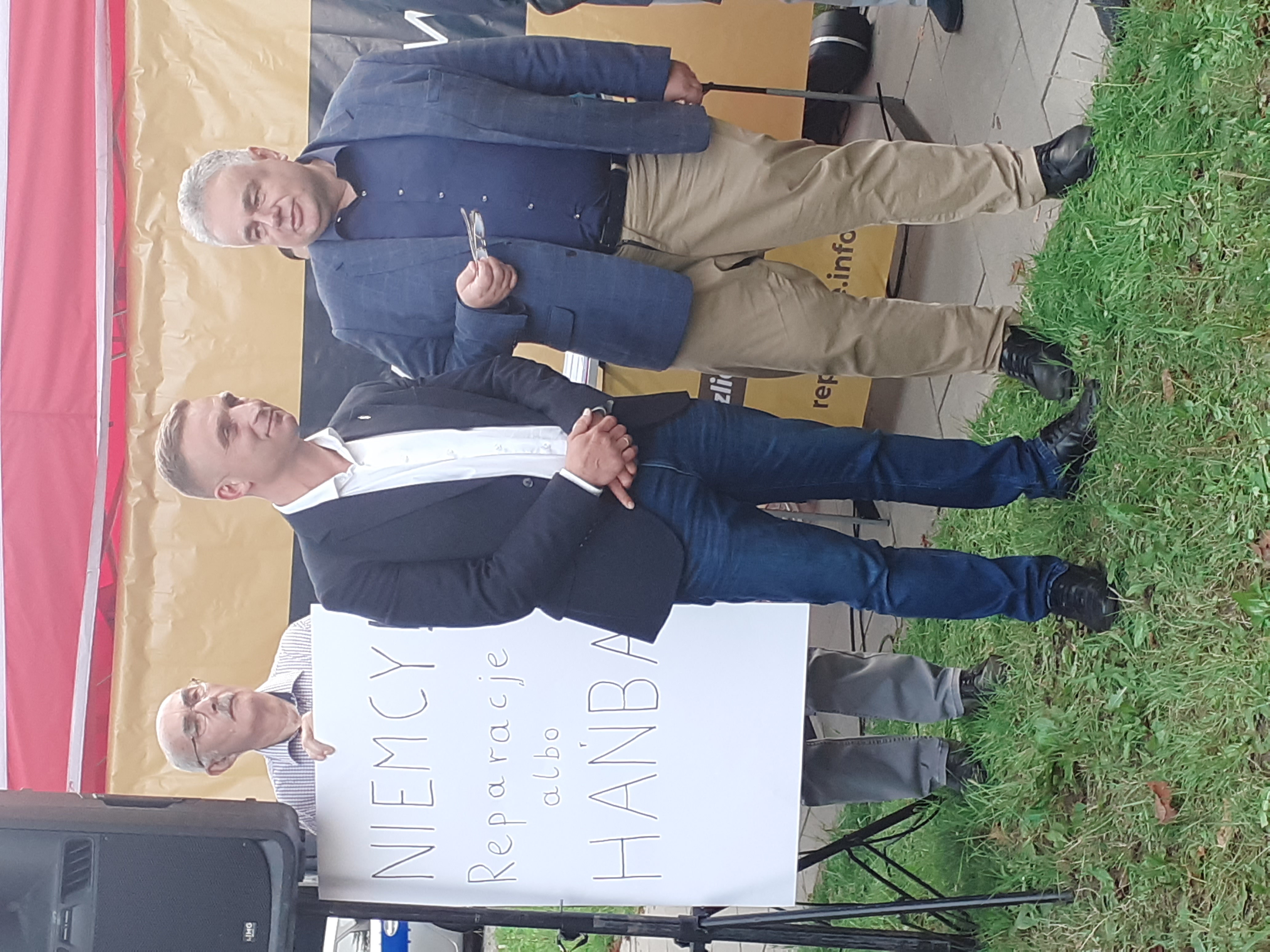 Tomasz Sakiewicz i inni mężczyźni na tle banera z napisem „NIEMCY Reparacje albo HANBA”