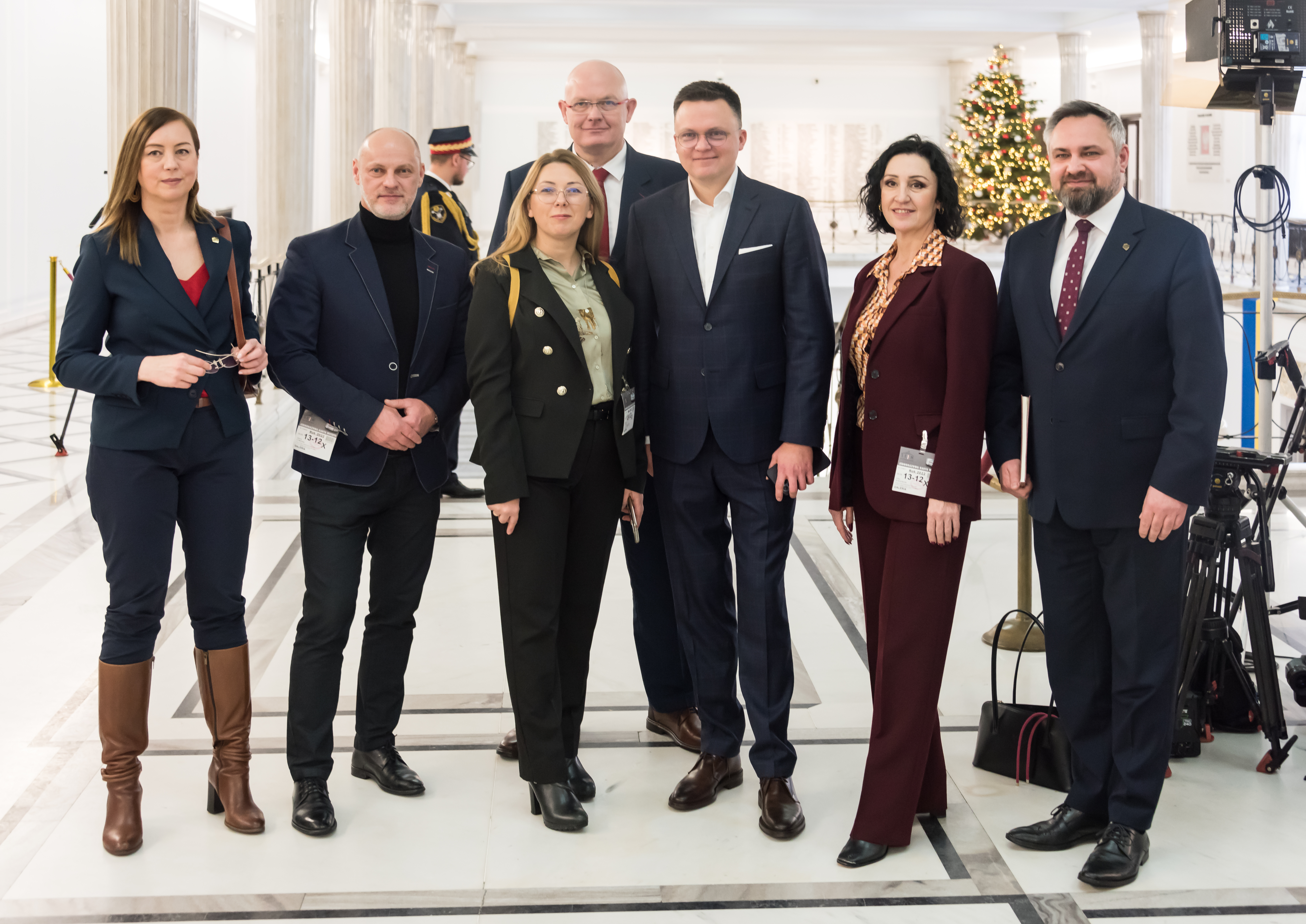 Grupa osób w eleganckich strojach, w tym Szymon Hołownia, przed choinką i sprzętem medialnym.