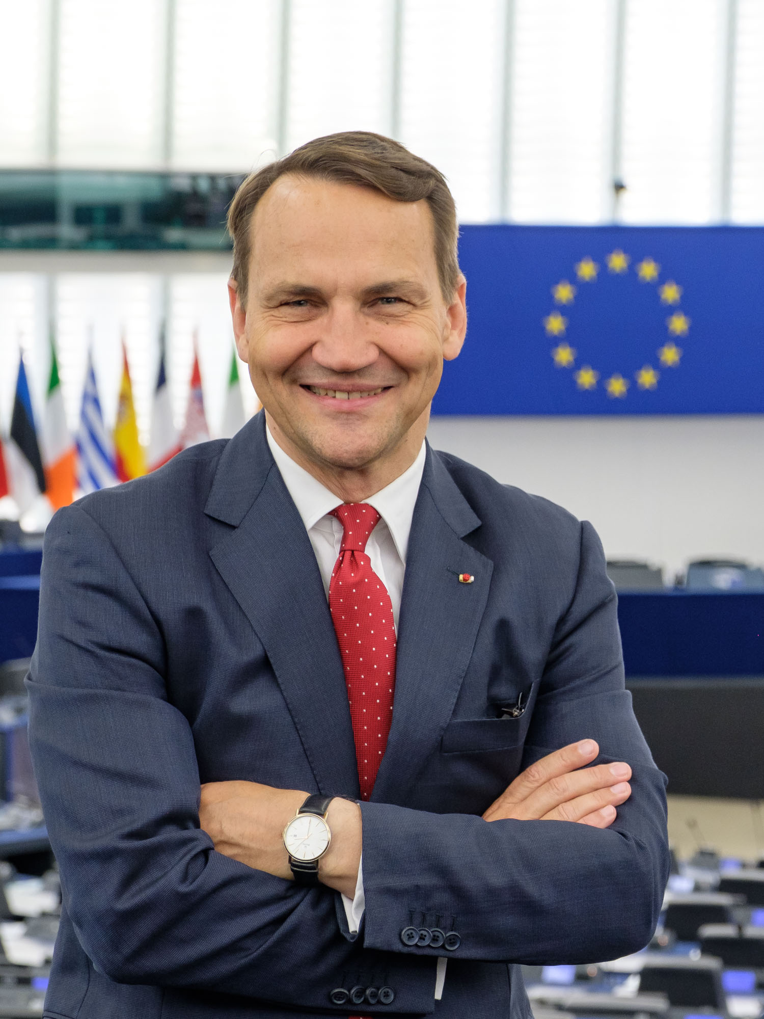 Radosław Sikorski w garniturze z krawatem, na tle flag Unii Europejskiej.