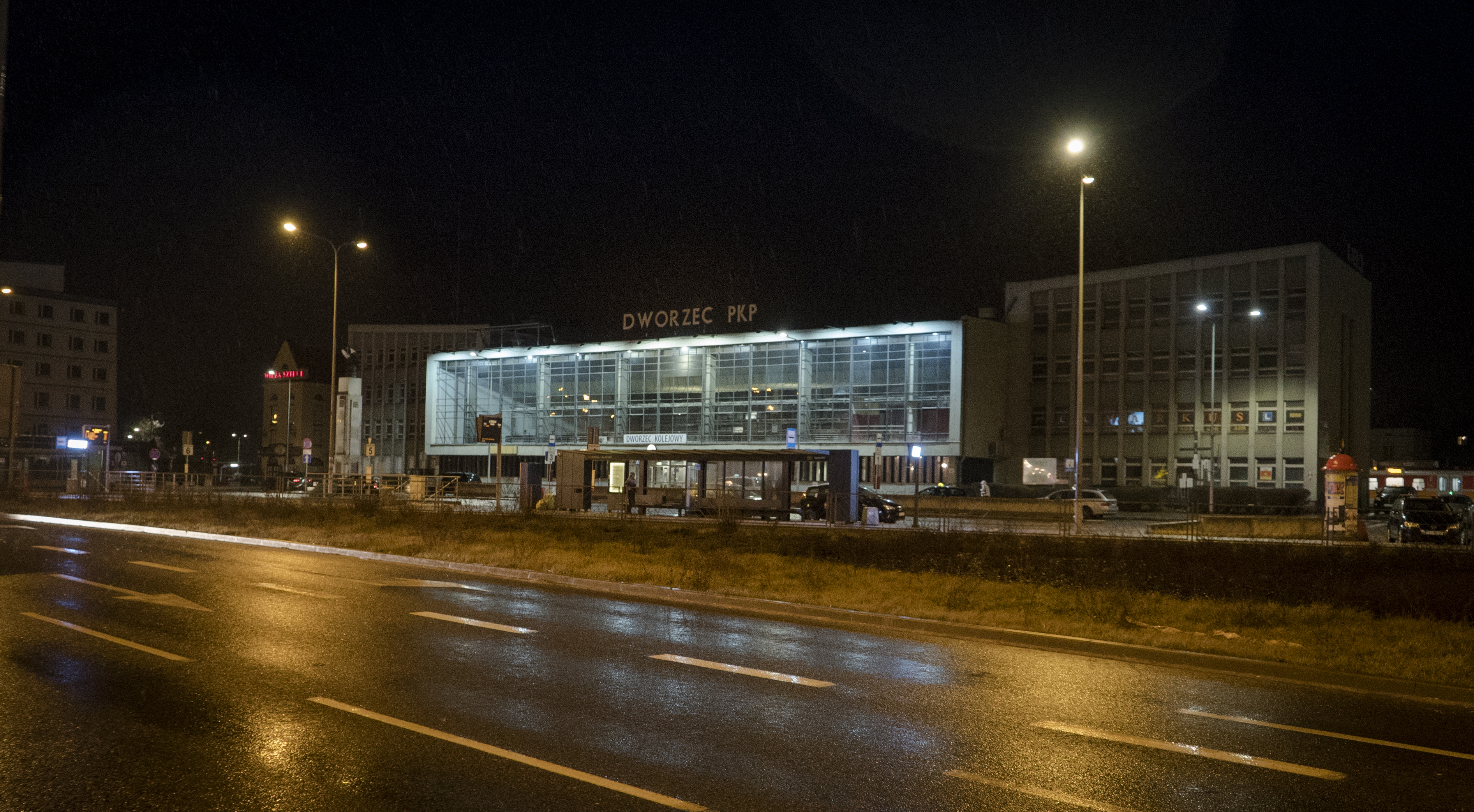 Budynek dworca PKP w Kielcach nocą, z widocznym deszczem i mokrą nawierzchnią drogi.