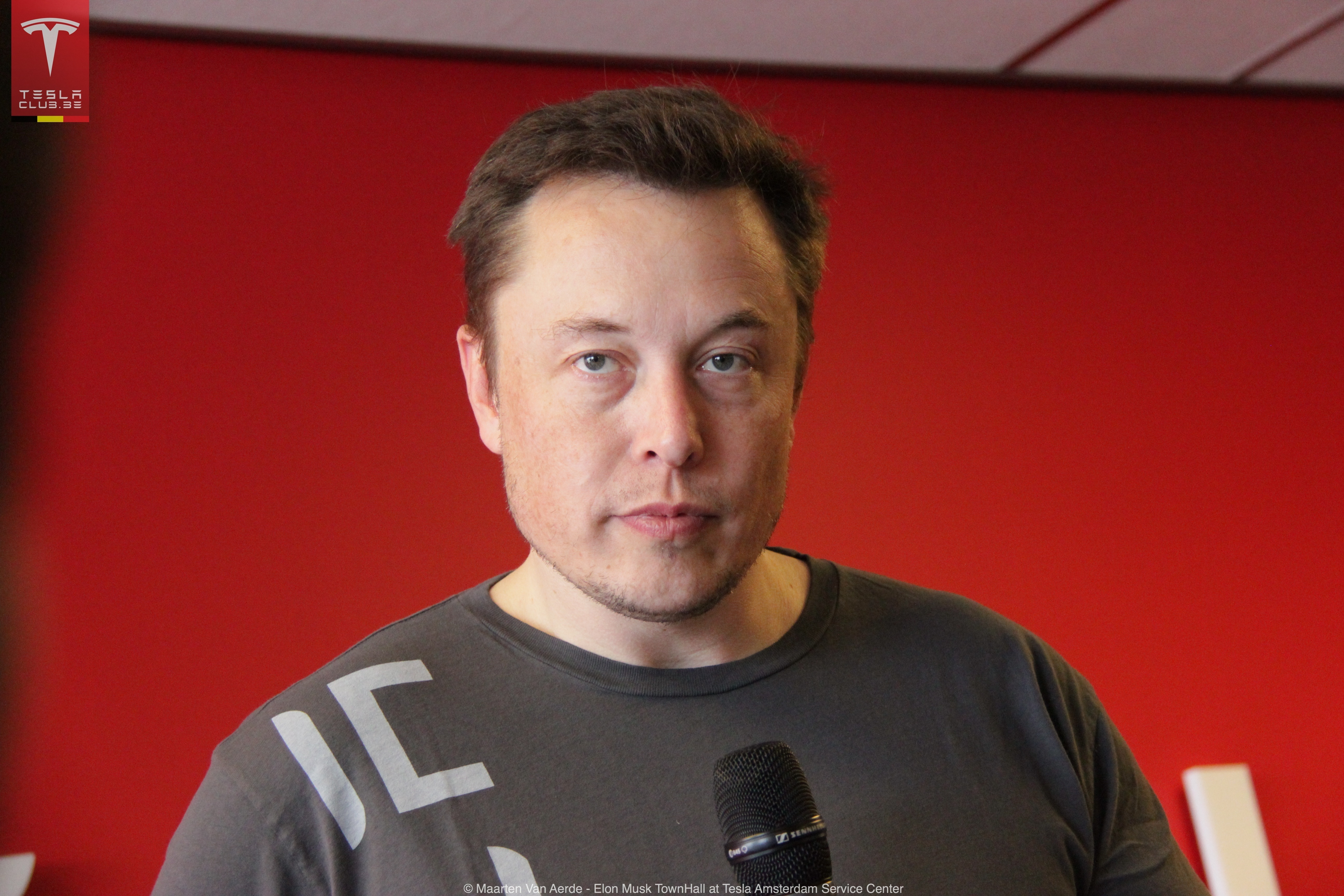 Elon Musk w koszulce z logo Tesla, przed czerwonym tłem.