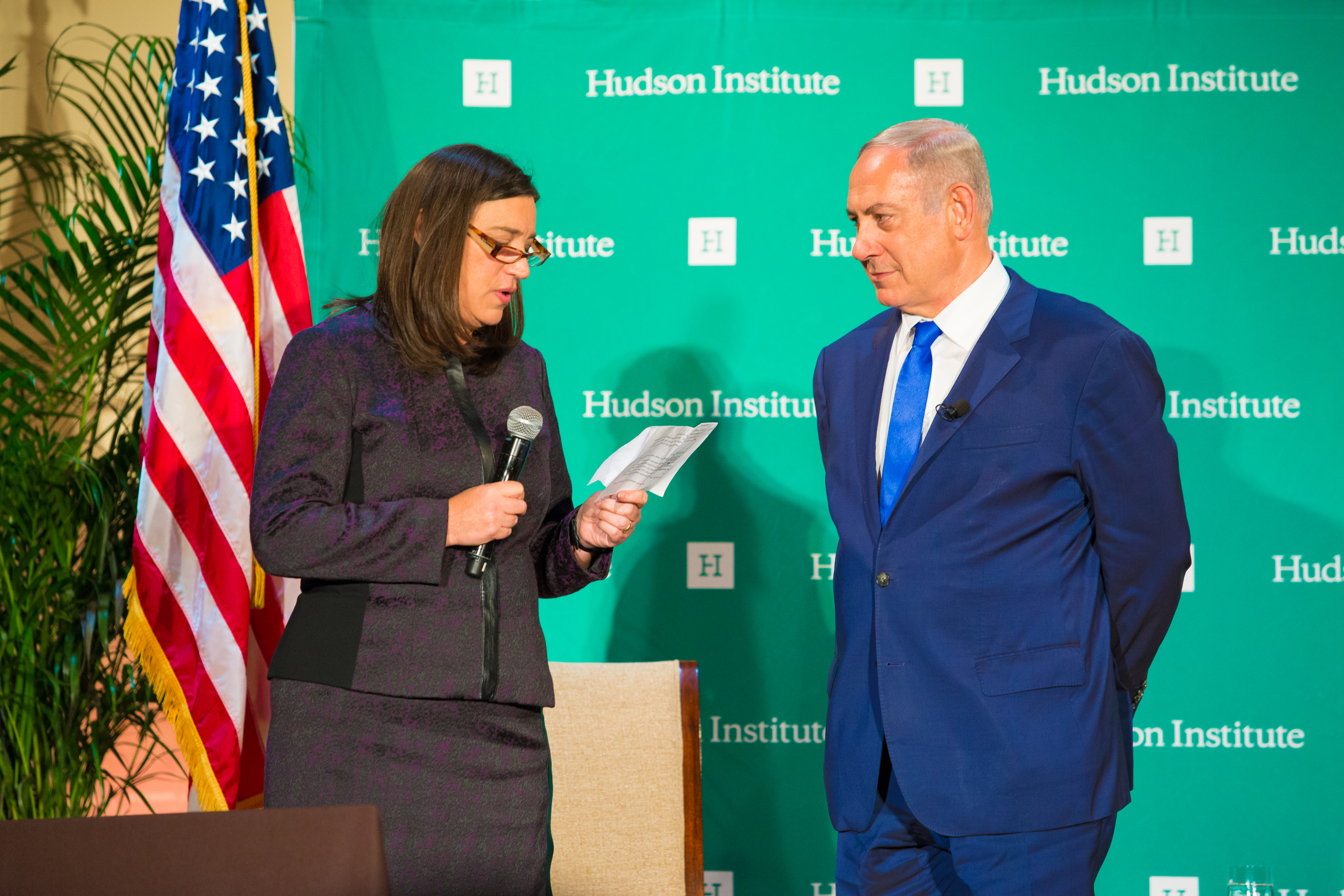 Binjamin Netanjahu w garniturze i kobieta z mikrofonem przed flagą USA i logo Hudson Institute.