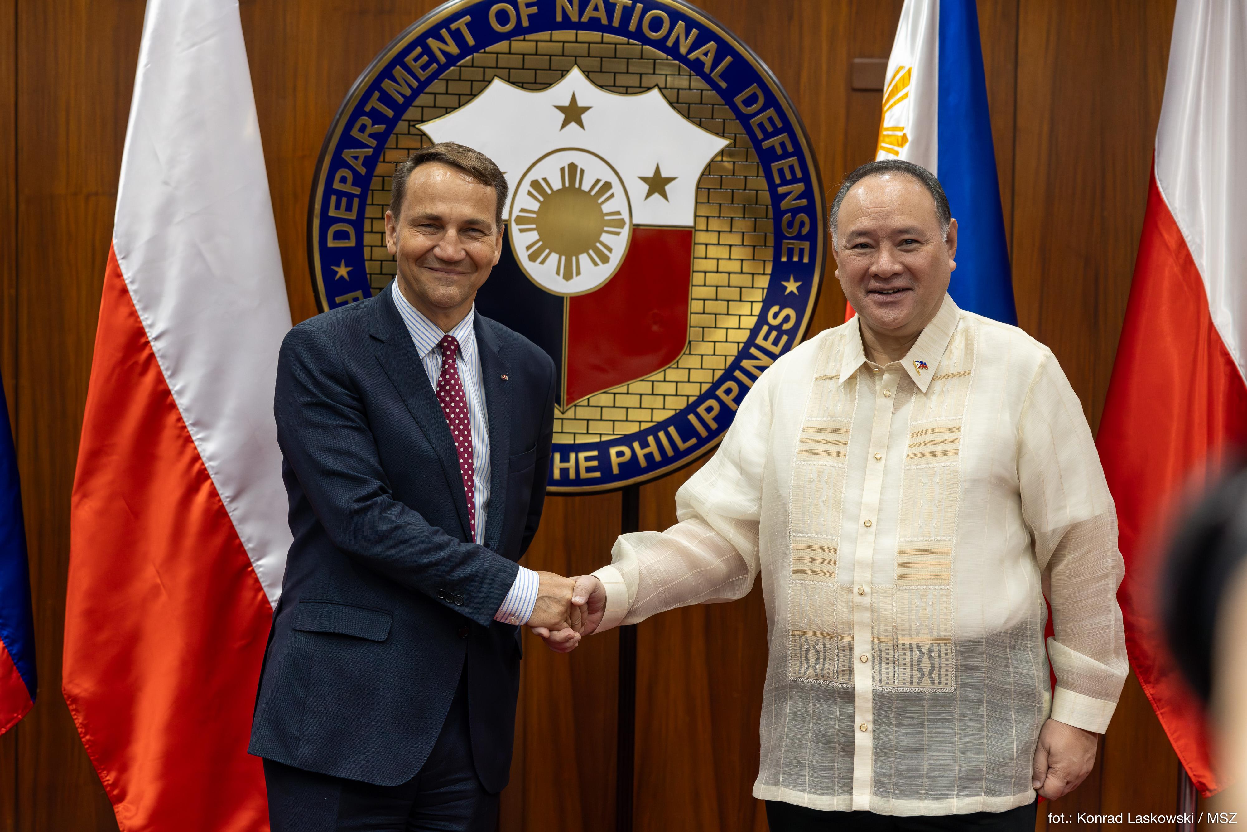 Radosław Sikorski wita się z przedstawicielem Filipin na tle flag i godła Departamentu Obrony Narodowej Filipin.