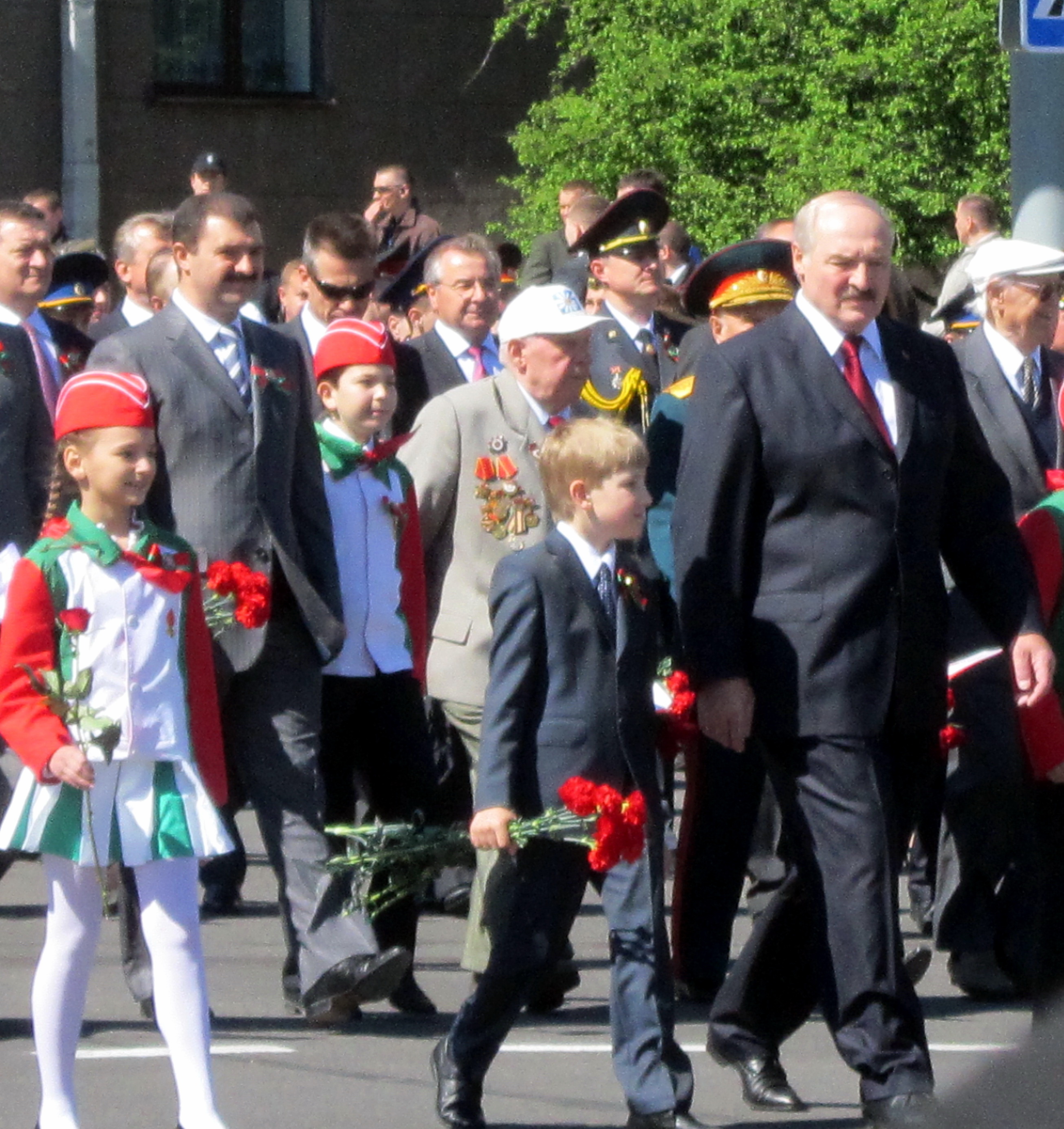 Aleksandr Łukaszenka w garniturze z czerwoną różą, podczas przemarszu z dziećmi na oficjalnym wydarzeniu.