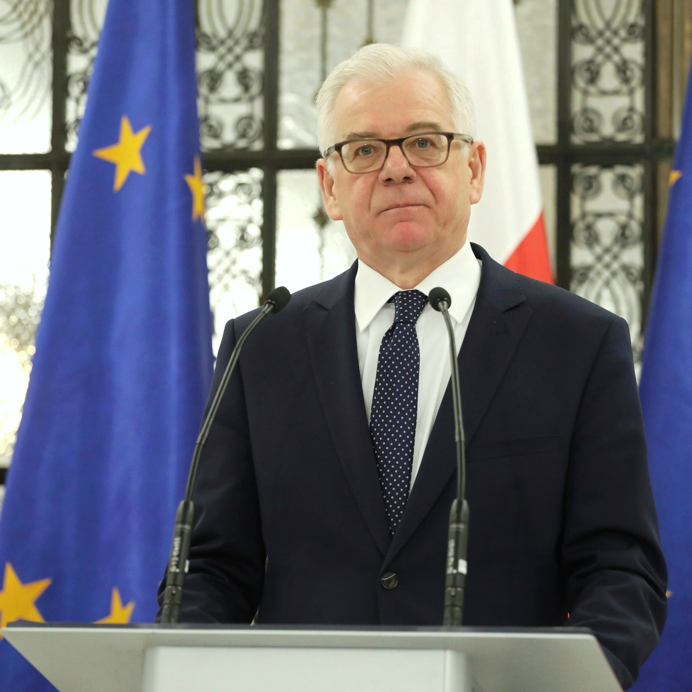Jacek Czaputowicz, były szef MSZ, przemawia na tle flagi Polski i UE, analizując bezpieczeństwo Polski.