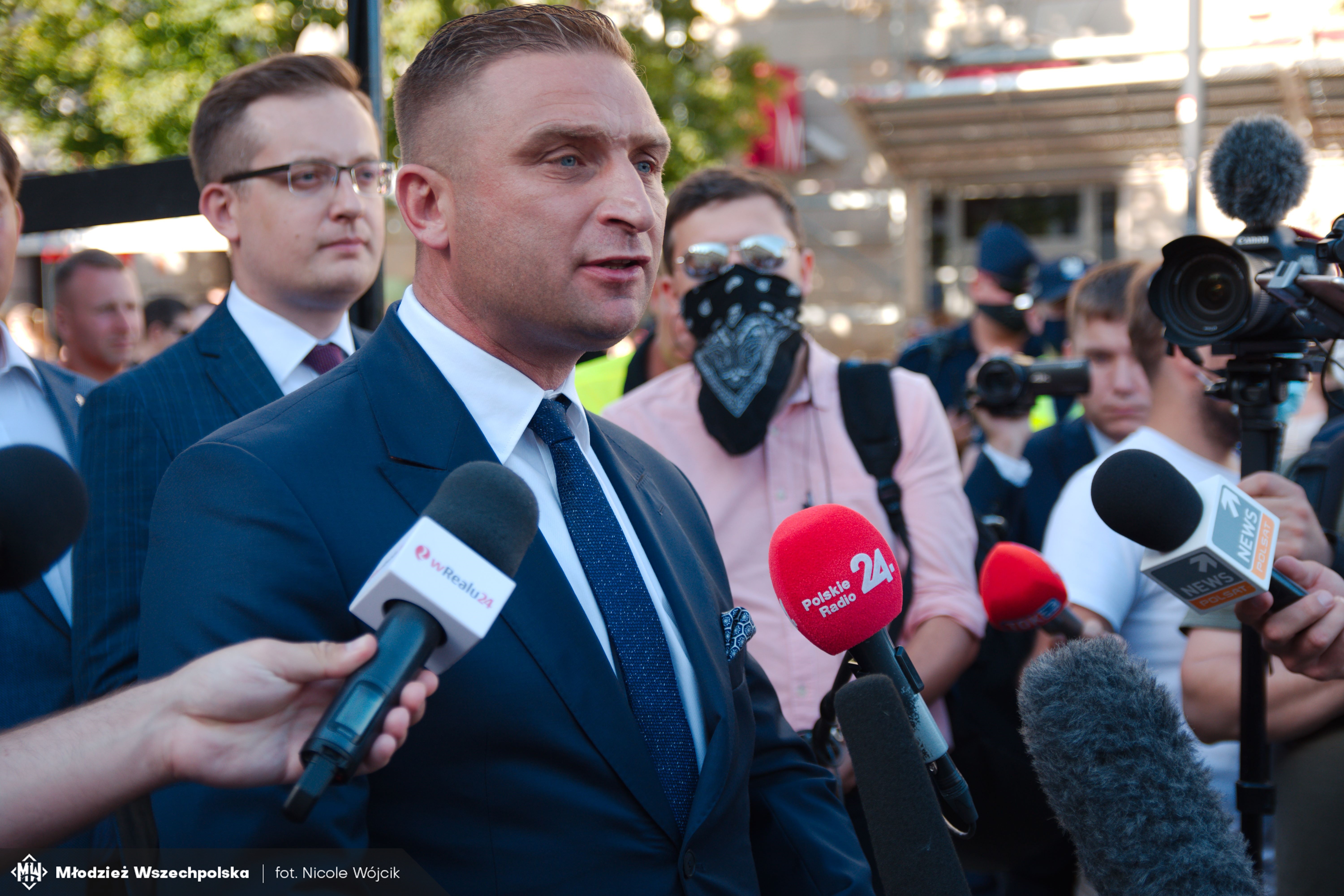 Robert Bąkiewicz udziela wywiadu dziennikarzom, przed nim mikrofony.