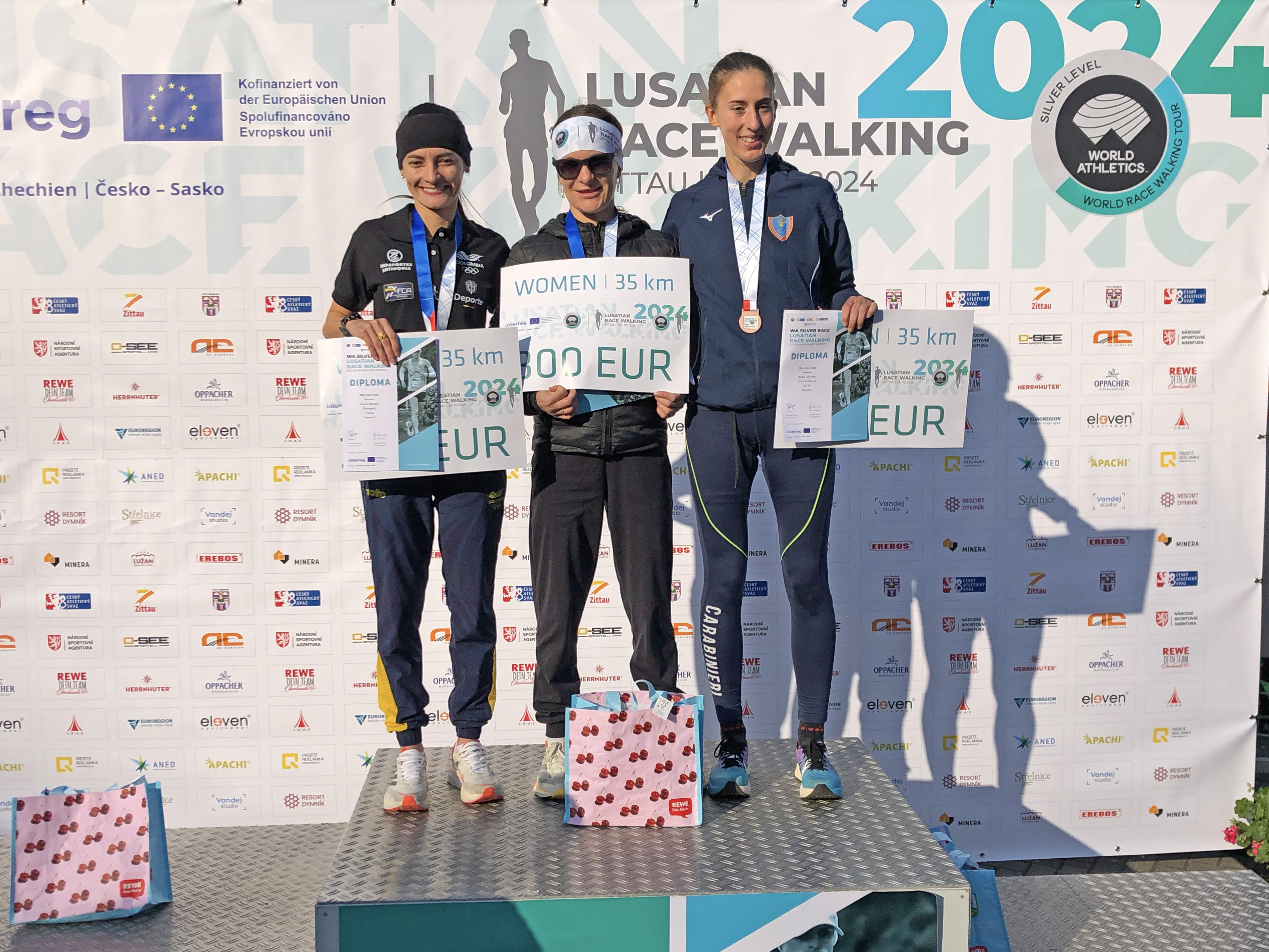 Kobiety na podium zawodów lekkoatletycznych w chodzie sportowym, z dyplomami i medalami.