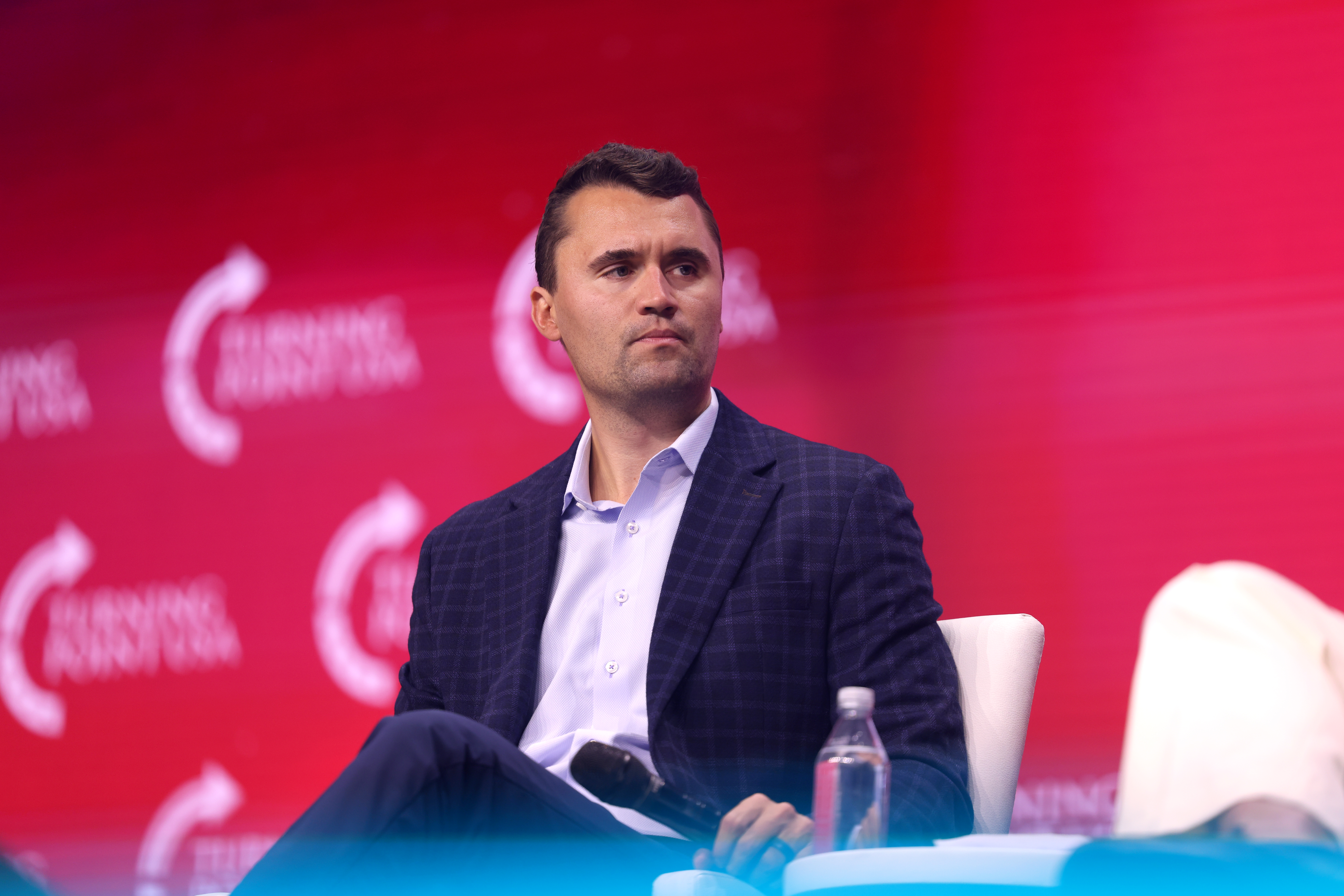 Charlie Kirk na scenie podczas wydarzenia Turning Point USA, siedzi z mikrofonem w dłoni.