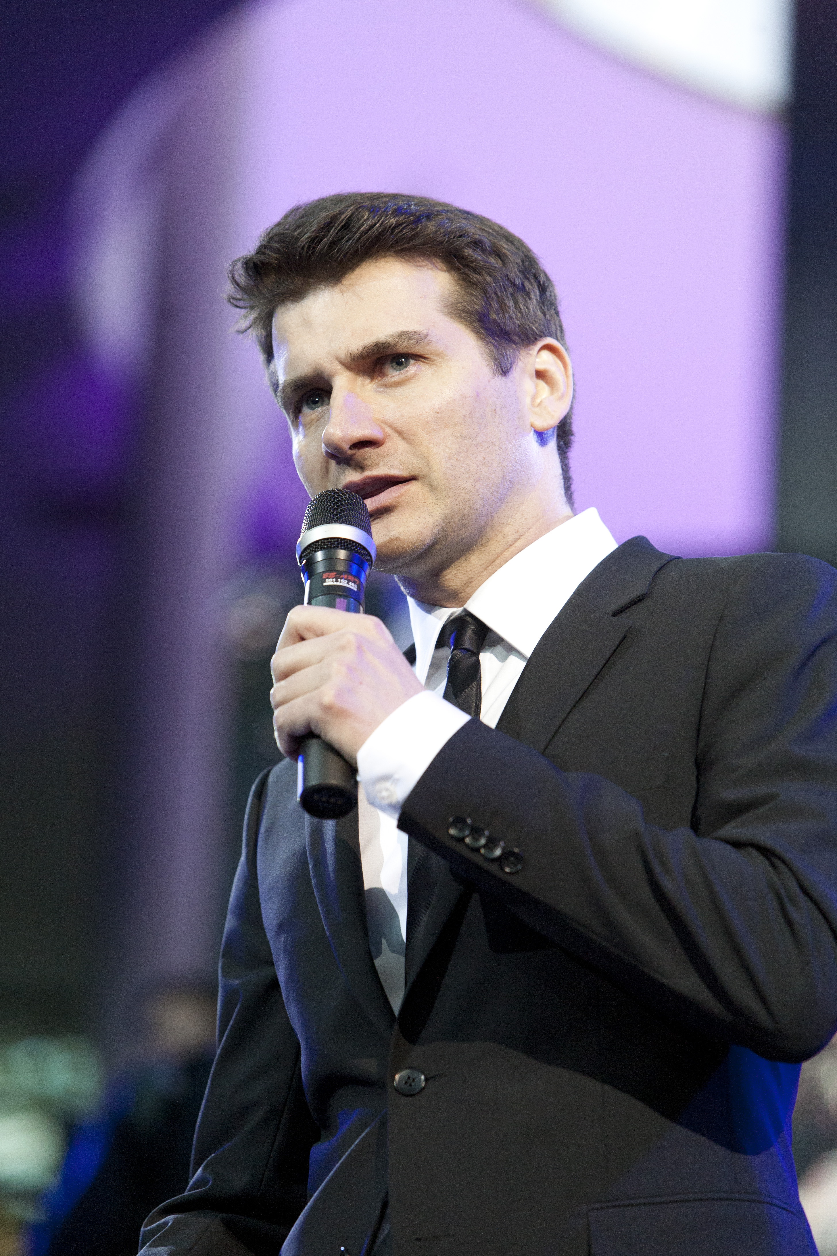 Aktor Tomasz wypowiada się do mikrofonu w garniturze.