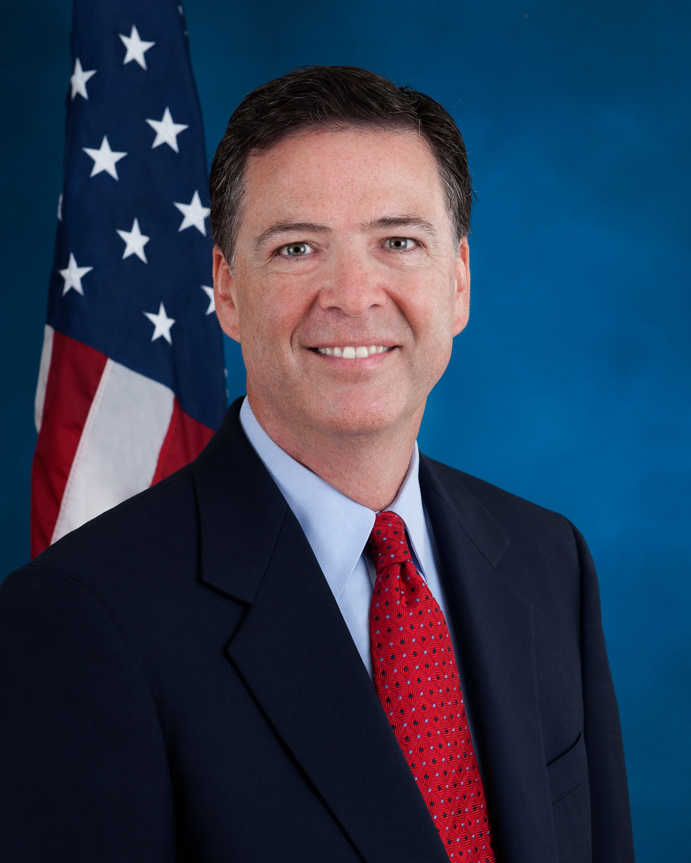 James Comey, były szef FBI, w garniturze i krawacie, na tle flagi USA.