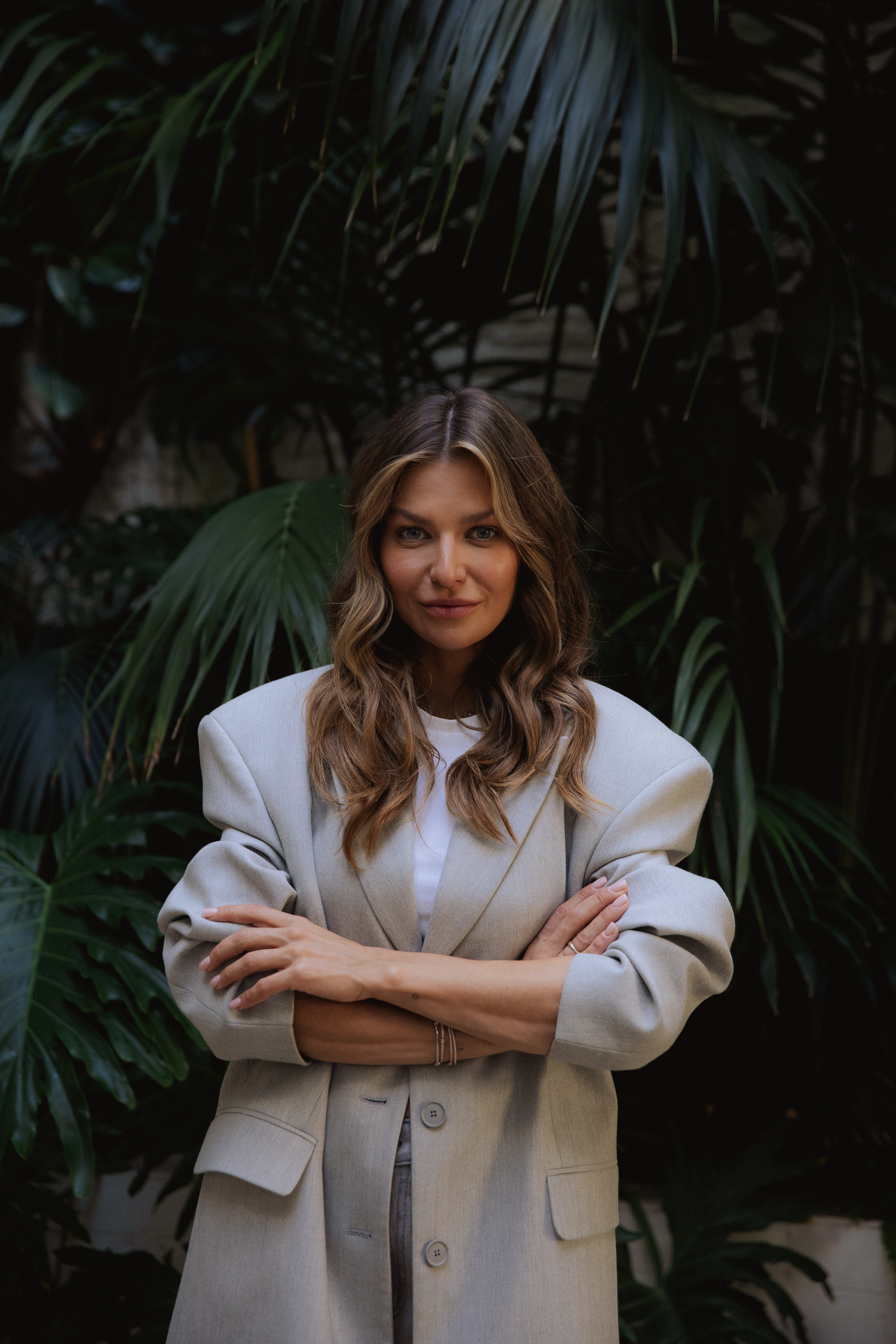 Anna Lewandowska w eleganckim stroju, z ramionami skrzyżowanymi na piersi, na tle zielonych liści.