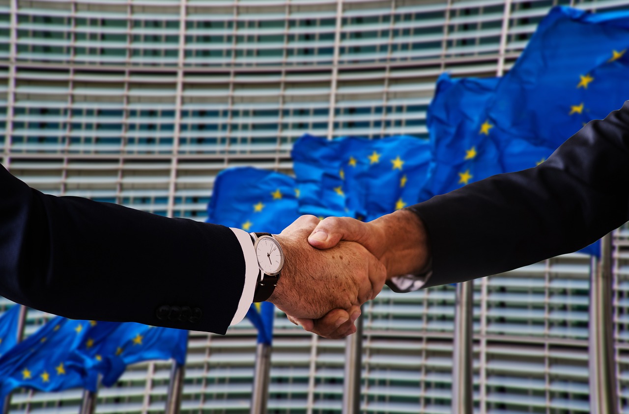 Dwie osoby ściskają sobie dłonie na tle flag Unii Europejskiej, symbolizujące negocjacje.