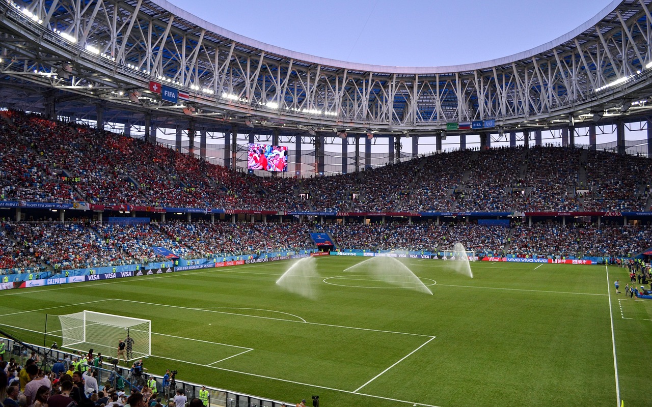 Stadion piłkarski z trybunami wypełnionymi kibicami, boisko z bramką i zraszaczami.