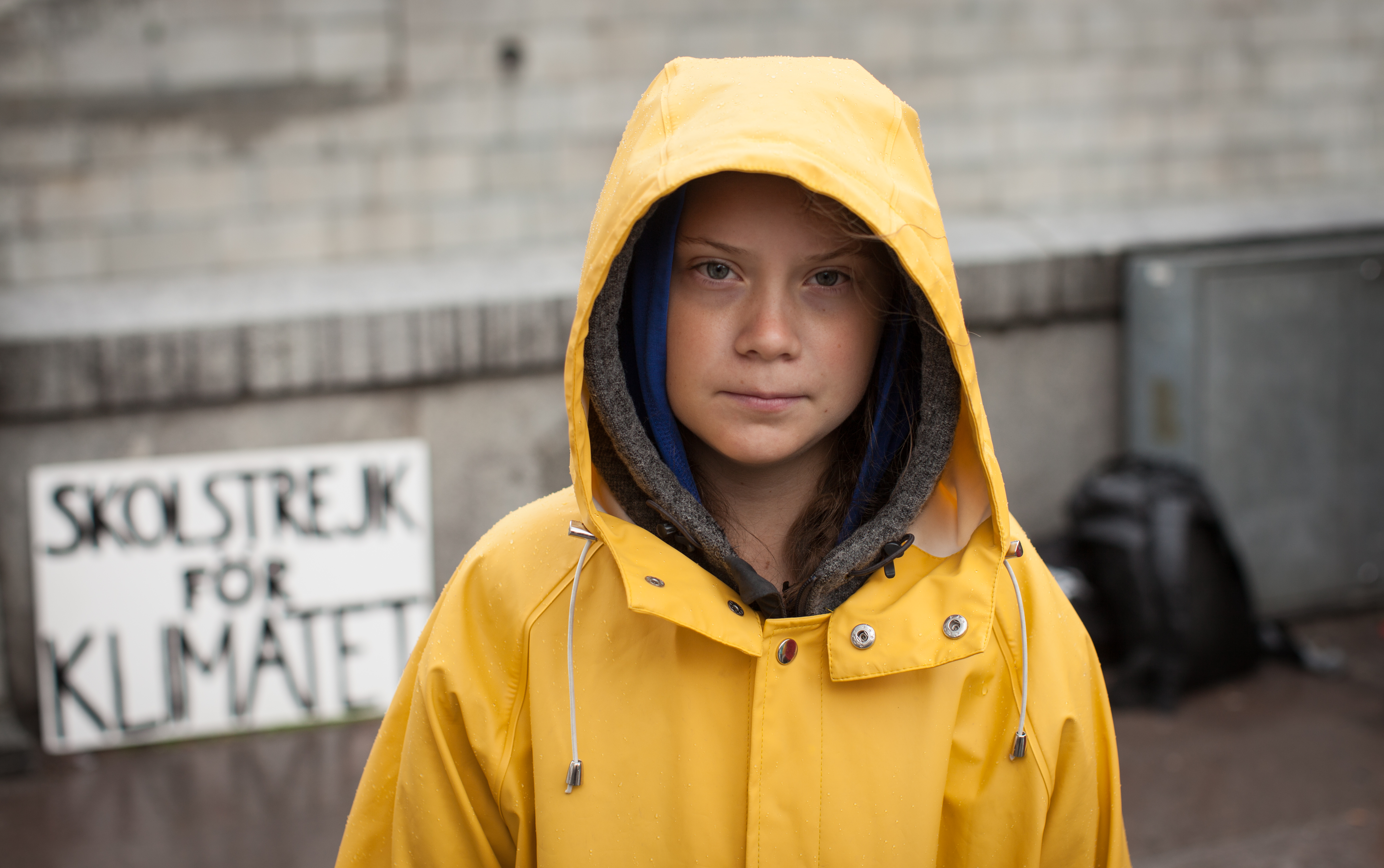 Greta Thunberg w żółtej kurtce przeciwdeszczowej z napisem "SKOLSTREJK FOR KLIMATET" w tle.