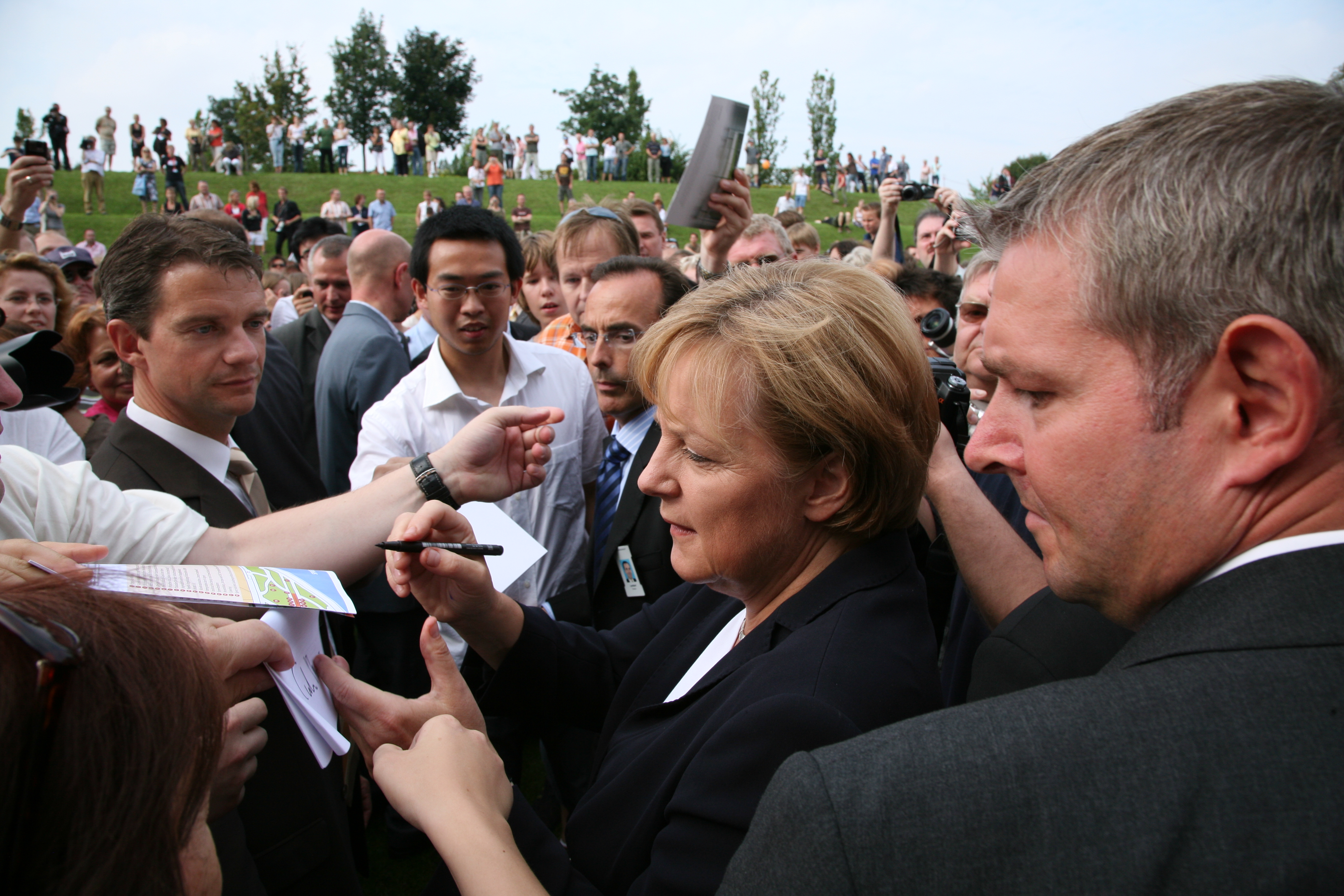 Angela Merkel podpisuje dokument, otoczona przez ludzi i dziennikarzy.