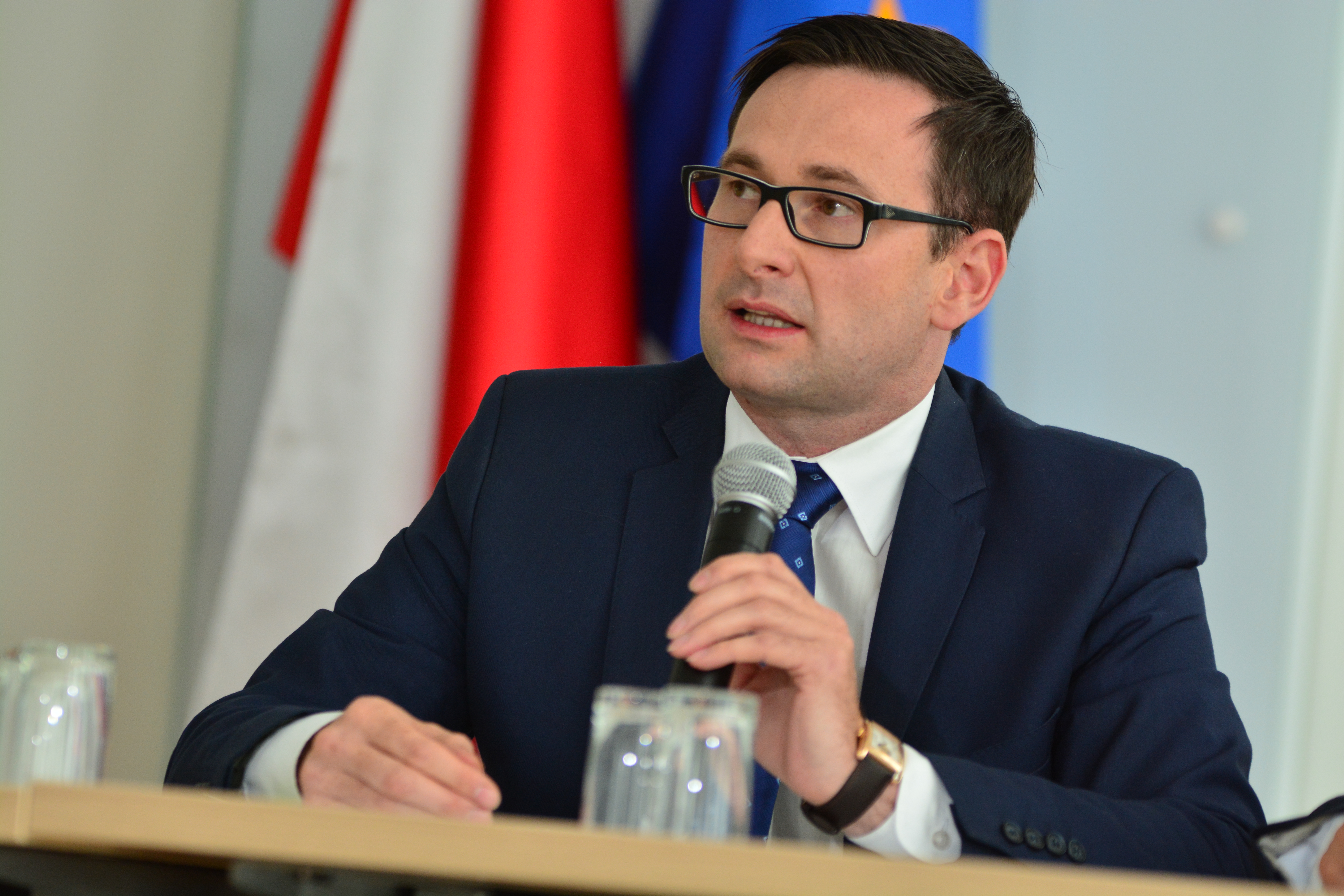 Daniel Obajtek w garniturze i okularach, trzymający mikrofon, podczas konferencji.
